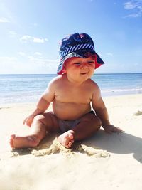 Shirtless baby sitting at beach on sunny day against sky