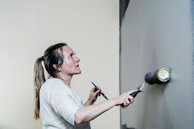 Woman catching with a hand a paint roller full of grey painting and with the other a brush. she is upping and downing the roller covering the wall with grey painting what remains wet. horizon