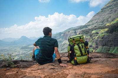 Rear view of man sitting by backpack on mountain against sky