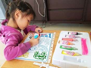 High angle view of girl playing with toy sitting on table and squeezing painting glue on the paper 