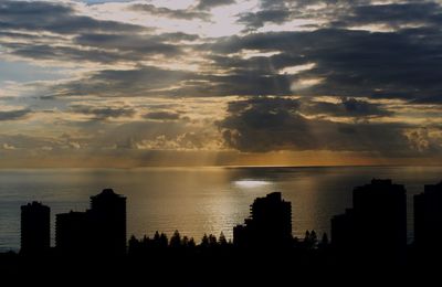 Silhouette buildings by sea against sky during sunset