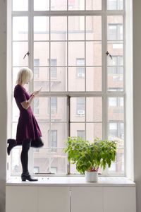 Side view of mature woman using mobile phone while standing on window sill