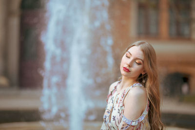 Beautiful young woman with eyes closed standing against fountain in city