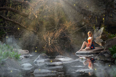 Young woman sitting by lake against trees