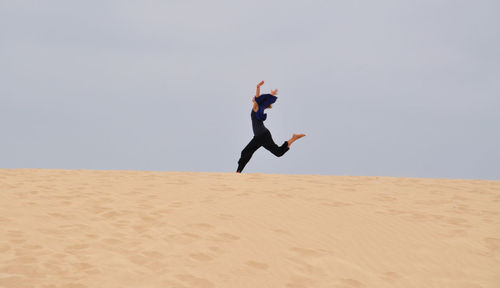 Cheerful woman running with arms raised on sand against clear sky at beach