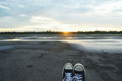 Low section of man sitting on road against cloudy sky during sunset