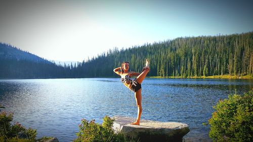Full length of woman practicing yoga on rock by lake against sky