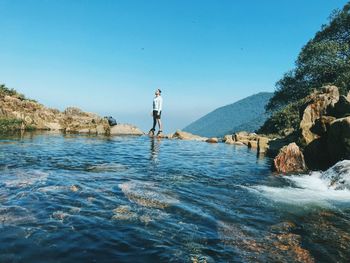 Young man standing on rock by lake against clear blue sky