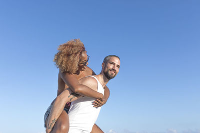 Low angle view of man piggybacking cheerful girlfriend against blue sky