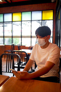 A handsome young teenage boy wear mask playing on smart mobile phone waiting in cafeteria. 