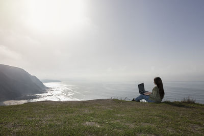 Rear view of woman sitting on rock by sea against sky