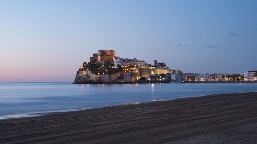 Sea by illuminated buildings against sky at dusk