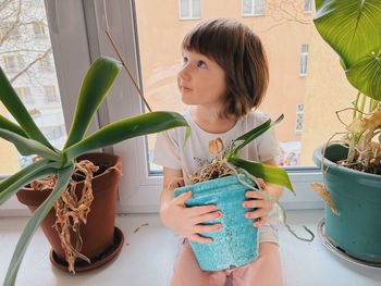 A toddler girl sitting at the window between house plants vwhile curiously looking at something