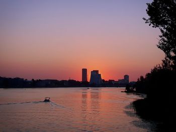 Silhouette buildings by trees against sky during sunset