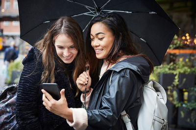 Smiling female friends with umbrella looking at mobile phone while standing in city