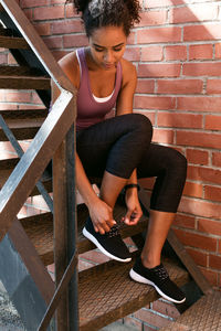 Full length of woman tying shoe laces while sitting on steps by brick wall