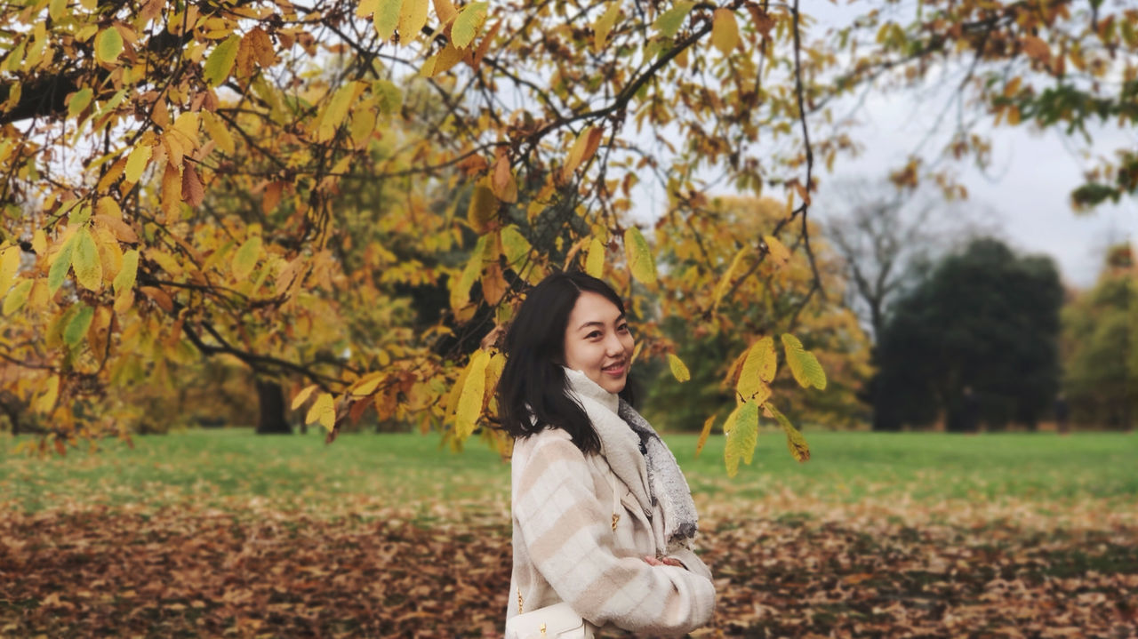 HAPPY YOUNG WOMAN STANDING BY TREE ON AUTUMN LEAVES