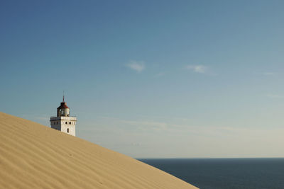 Sand dune by lighthouse at shore against sky