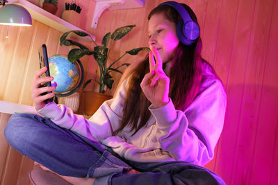 Happy cute pretty teen in headphones joying, in neon room at home. girl making selfie, listen music