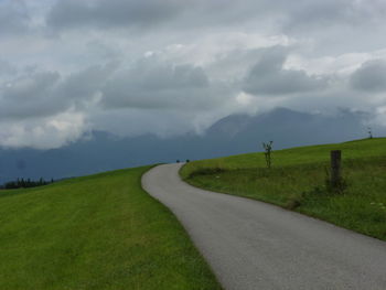 Road passing through grassy field against cloudy sky