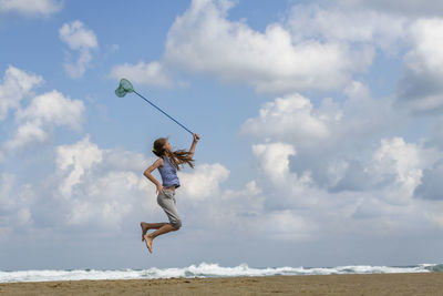 Full length of girl holding butterfly net while jumping on beach against sky