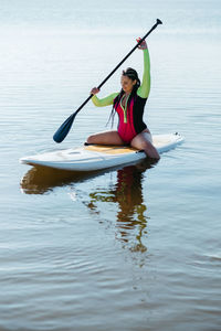 Young woman with dreadlocks in swimwear sitting on the sup board on the lake at sunrise