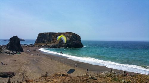 Scenic view of paraglidier over sea and beach against arched rock and clear sky