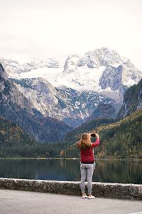 Rear view of woman standing on mountain by lake taking a picture