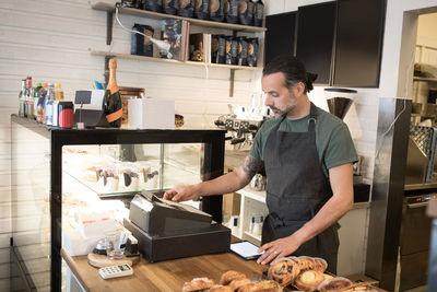 Confident mature male owner using cash register at checkout in bakery