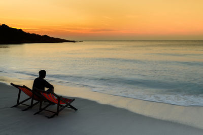 Man sitting on seat at beach against sky during sunset