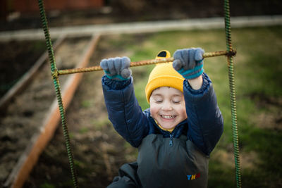 Cute smiling boy wearing warm clothing climbing on rope ladder at playground