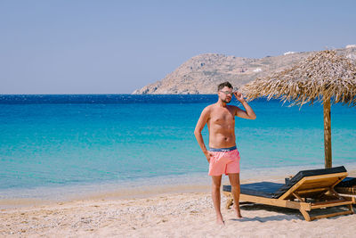 Full length of shirtless man standing on beach against clear sky