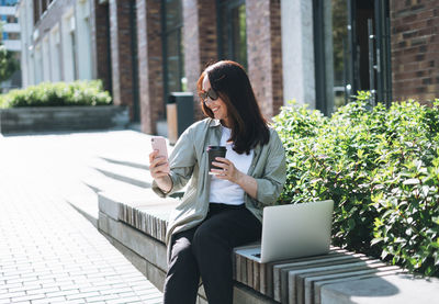 Adult woman forty years in stylish shirt working on laptop and using mobile phone on bench 