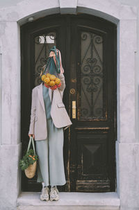Woman covering face with mesh bag full of oranges in front of door