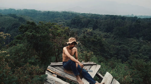 Full length of shirtless man sitting on wood against mountains