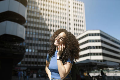 Smiling woman in eyeglasses talking on smart phone near building