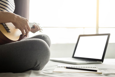 Midsection of woman playing guitar while sitting by laptop on bed