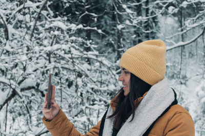 Side view of girl wearing winter clothes, using mobile phone and taking photos in snowy forest