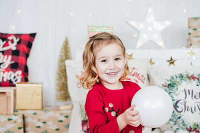 Happy little girl wearing red christmas dress at home over christmas decoration. holding bauble