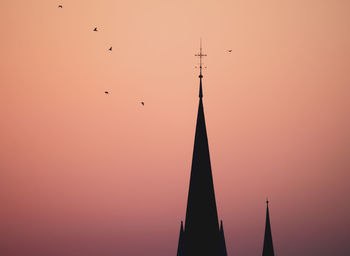 Low angle view of silhouette of birds and church towers against sky during sunset