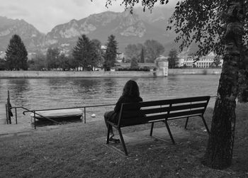 Rear view of woman sitting on bench by lake against town
