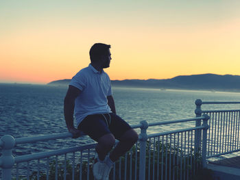 Full length of man sitting on railing against sea during sunset