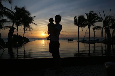 Silhouette people standing by swimming pool against sky during sunset