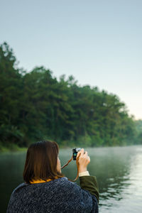 Rear view of woman photographing lake against trees