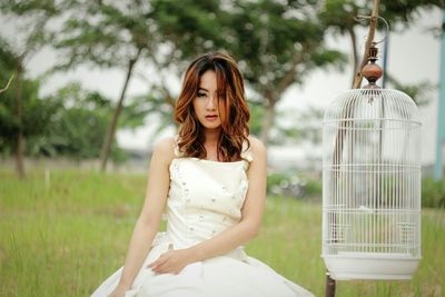 Portrait of young woman sitting by birdcage over field