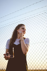 Young woman drinking glass while standing by fence against sky