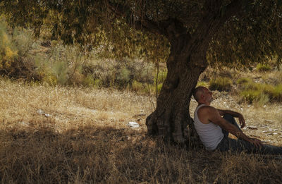 Adult man in white tank top and jeans taking rest under olive tree in summer