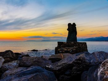 Statue on rock at beach against sky during sunset