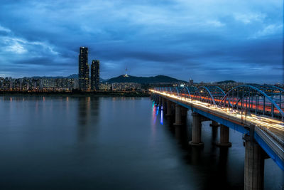 Illuminated bridge over river against cloudy sky