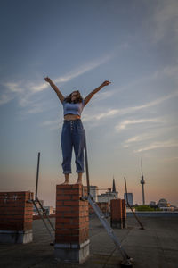 Happy woman with arms raised standing on rooftop against sky during sunset